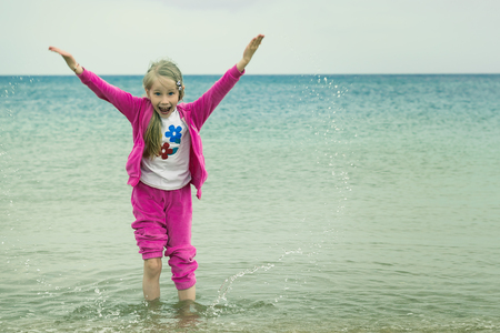 Cheerful girl in a warm suit plays with water in a cold sea. The concept of a happy childhood on the ocean.の写真素材