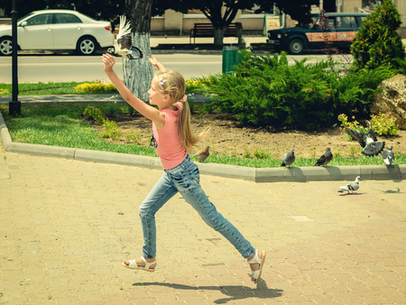 Girl in a summer day catching a flying dove. The child plays with birds in the town square.の写真素材