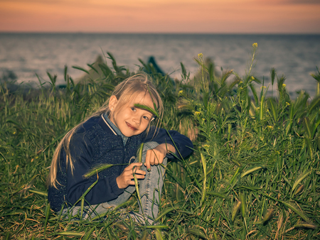 The girl hiding in the green grass on the beach at sunset. Portrait of a little beautiful girls in the grass by the sea.の写真素材