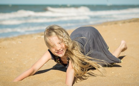 The girl in the gray dress on the sand by the ocean. Portrait of cheerful girls playing with sand and wind from the sea.の写真素材