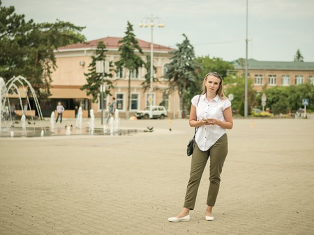 Young beautiful woman in the town square communicates through an electronic device. Portrait of woman in full growth in the city.の写真素材