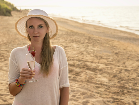 Beautiful woman in hat with glass of wine on the beach by the sea. Portrait of a woman with sparkling wine at the sea.の写真素材