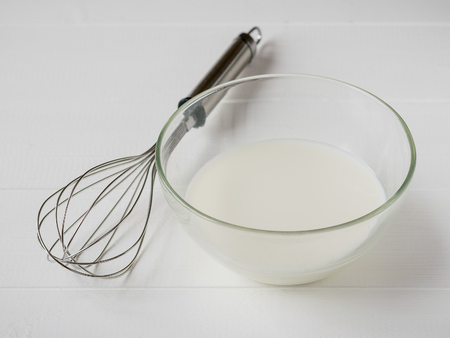 Whisk and glass bowl with milk on white wooden table. The ingredients for making scrambled eggs or baking.の写真素材