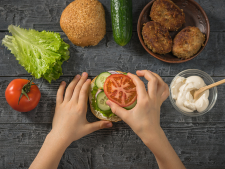 The girl puts a piece of tomato on a Burger. Cooking hamburger for school lunch child.の写真素材