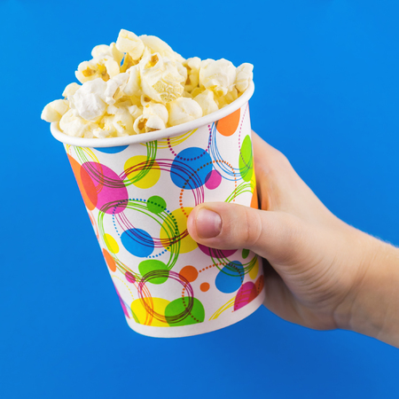 Children's hand with a full glass of popcorn on a blue background. The concept of partying and watching of movies.の写真素材