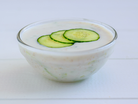 Three slices of cucumber in a bowl with tzatziki sauce on white wooden table.の写真素材