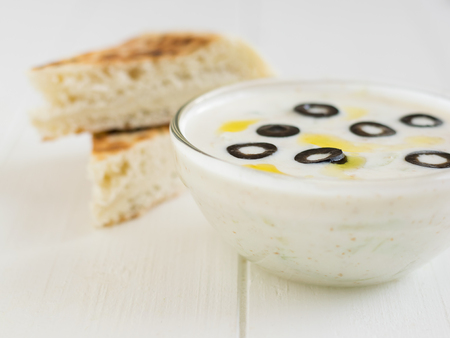 Two pieces of bread, and tzatziki in a bowl with olives on a white wooden table.の写真素材