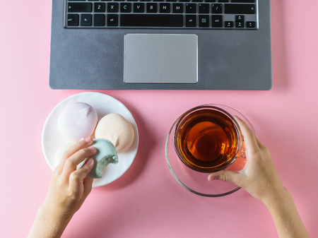 The girl eats a blue marshmallow with tea in front of the computer on a pink table. The view from the top. The concept of healthy eating in the modern world.の写真素材