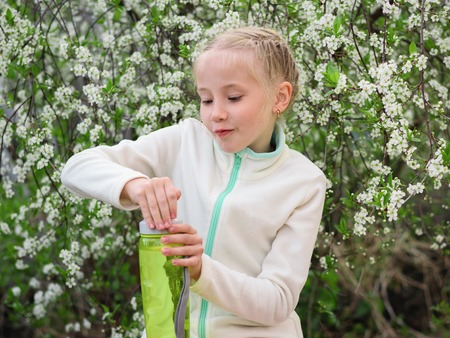 A beautiful girl in the spring Park quenches her thirst with water with lemon and mint. Portrait of a girl on a background of cherry blossoms.の写真素材