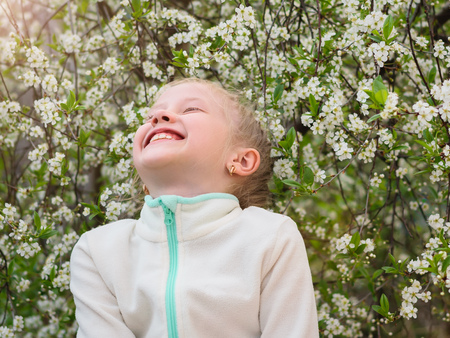 A girl in a sports jacket laughs merrily in a spring cherry Park. Portrait of a girl on a background of cherry blossoms.の写真素材