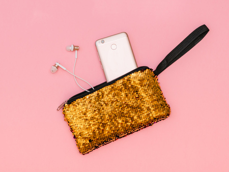 Women's handbag with a sticking phone and headphones of gold color on a pink table. Pastel color. Women's accessories. Flat lay.の写真素材