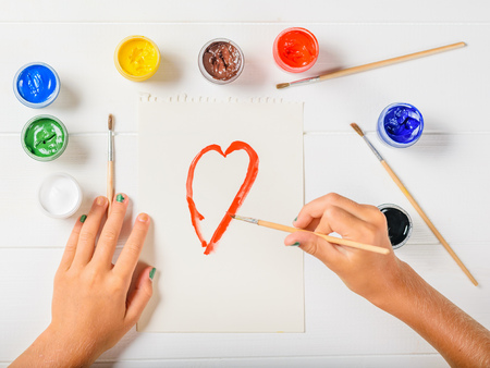The girl paints a red heart on a sheet of paper on a white wooden table. Creative kit. Hobby and style. Flat lay.の写真素材