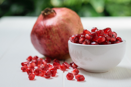 White Cup with pomegranate seeds and ripe pomegranate on white wooden table. Harvest ripe pomegranate . Vegetarian food. Raw food.の写真素材