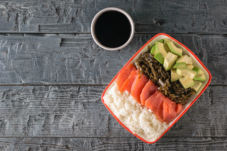 A plate of Hawaiian rice, avocado, salmon and kelp and soy sauce on a black wooden table....
