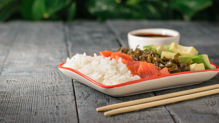 Bowl with rice, avocado, seaweed and salmon on a wooden table on a background of green vegetation. Mediterranean diet cuisine.の写真素材