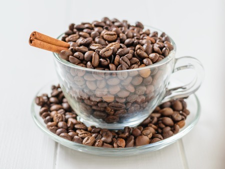 Glass bowl with coffee beans and cinnamon stick on white wooden table. Grains for the preparation of the popular drink.の写真素材