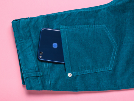 Blue phone sticking out of the pocket of green jeans on pink background. The concept of modern fashion for electronic devices.の写真素材