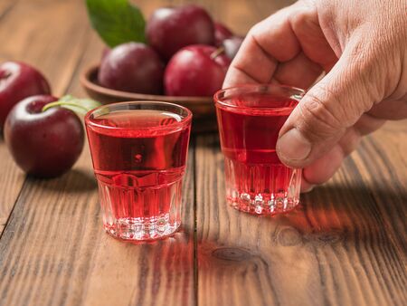 A glass of plum alcohol in a man's hand on a wooden table. Homemade alcoholic drink made from berries plum.の写真素材