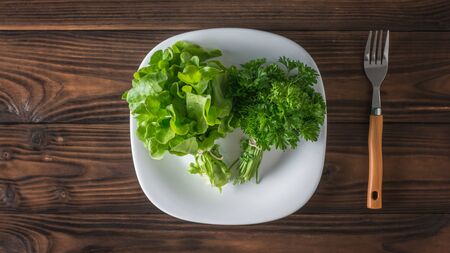 Fresh greens on a white plate and a fork on a wooden table. The concept of healthy eating. Flat lay.の写真素材