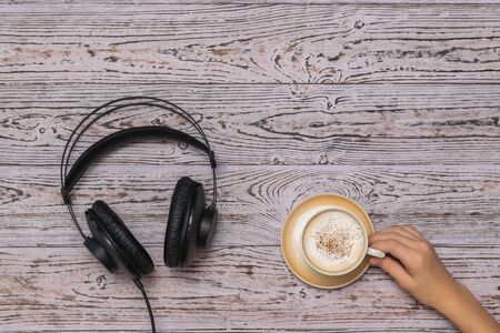 Hand holding a coffee Cup and black wired headphones on the wooden table. Coffee break during work.の写真素材