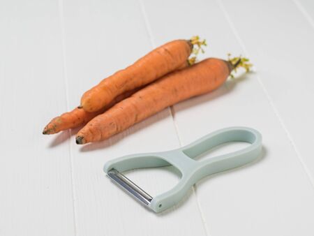 Three carrots and a peeler on a white rustic table. Cleaning carrots with a special knife.の写真素材