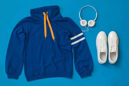 Sweatshirt, white sneakers and white headphones on a blue background. Sports lifestyle accessories.