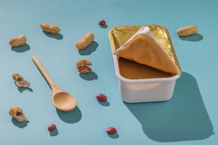 Peanut paste in a lunch box, wooden spoon and peeled peanuts on a blue background. Natural peanut...