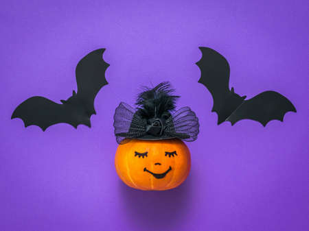 A pumpkin in a hat and two flying bats on a lilac background. The concept of the Halloween...