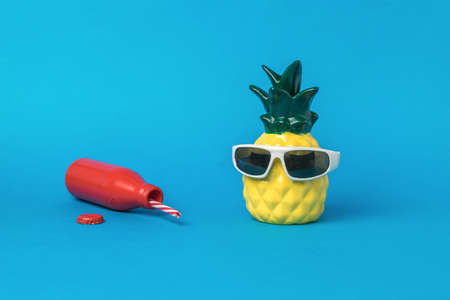 A pineapple figurine with sunglasses and a fallen red bottle on a blue background. Summer concept.の写真素材