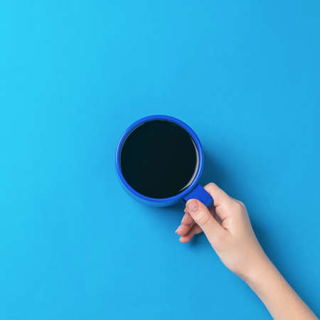 The girl's hand holds a blue glass with black coffee on a bright blue background. A woman's hand with a popular invigorating drink.の写真素材