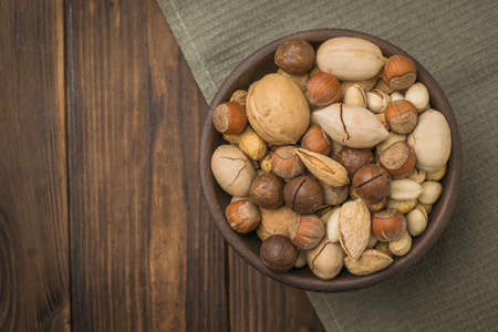 A bowl filled with a mixture of different nuts on a piece of cloth on a wooden table. Vegetarian...