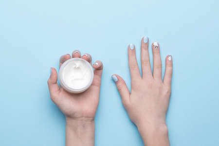 A jar of cream in women's hands with a stylish manicure on a blue background. Hand skin care. Flat lay.の写真素材