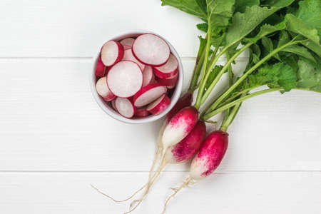 Top view of a bunch of radishes and a bowl of radish slices on a white wooden table. A fresh crop of...