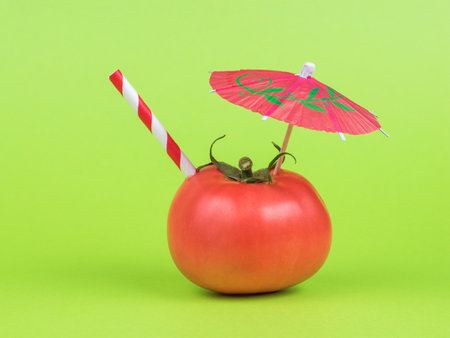 A red cocktail umbrella and a red tube in a tomato on a green background. The concept of consumption of fresh vegetable juice.の写真素材