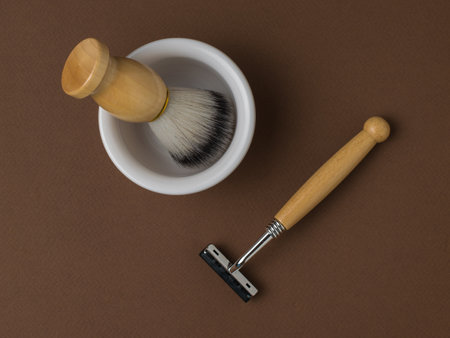 A brush in a white bowl and a razor with a wooden handle on a brown background. Men's accessories for appearance care. Flat lay.の写真素材