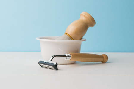 A bowl with a brush and a razor with a wooden handle on a white table on a blue background. Men's accessories for appearance care.の写真素材