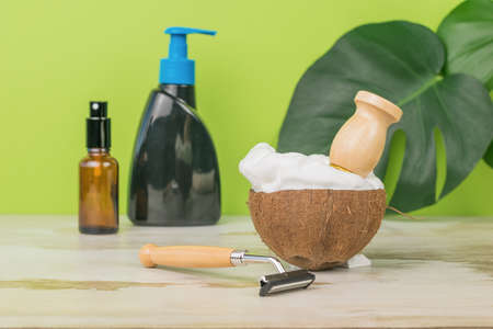 Coconut bowl filled with shaving foam and washing accessories on a green background with leaves. Men's accessories for appearance care.の写真素材
