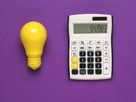 A calculator and a bright yellow light bulb on a purple background. The concept of increasing electricity payments.の写真素材