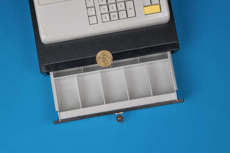An open empty cash register and a digital coin on a blue background. The concept of replacing money...