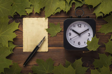 A sheet of old paper with a pen and a retro clock on a wooden background with leaves. Autumn concept. Flat lay.の写真素材