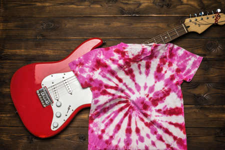 Red and white tie dye T-shirt and electric guitar on a dark wooden background. Staining fabric in tie dye style. Flat lay.の写真素材