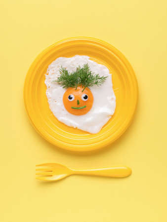 A face made of fried egg and parsley on a yellow plate and a yellow fork on a yellow background. minimal concept. A popular breakfast. flat lay.の写真素材