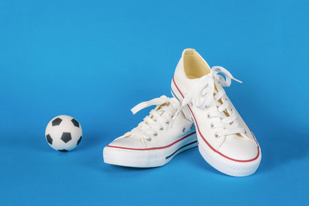 A pair of stylish white sneakers with laces and a small soccer ball on a blue background. Minimal sports concept.の写真素材