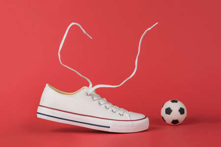 White sneakers with flying laces and a small soccer ball on a red background. Minimal sports concept.の写真素材