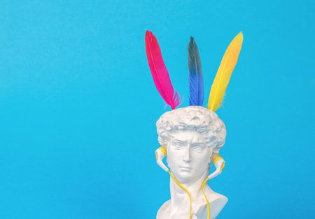 Plaster figure of Apollo with colorful bird feathers and headphones on a blue background. minimalism. Surrealism.の写真素材