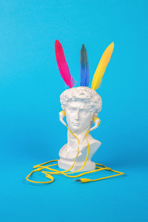 A statuette of Apollo wearing headphones with three colored bird feathers on a blue background. minimalism. Surrealism.の写真素材
