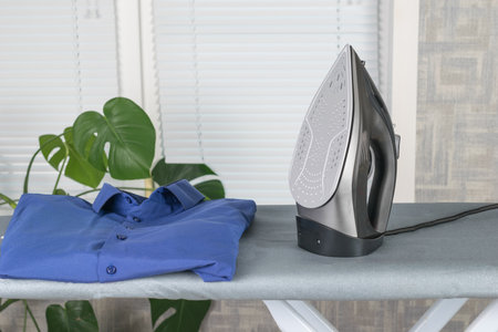 A blue men's shirt and an iron on an ironing board on the background of the window. Minimal concept of linen care.の写真素材