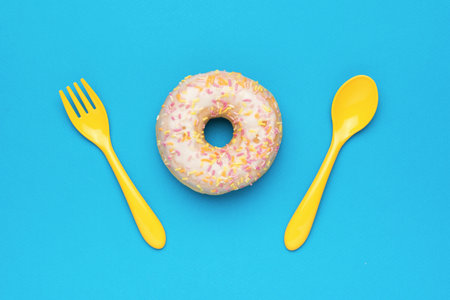 A yellow spoon and fork and a pink glazed donut on a blue background. The minimal concept of popular baking. flat lay.の写真素材
