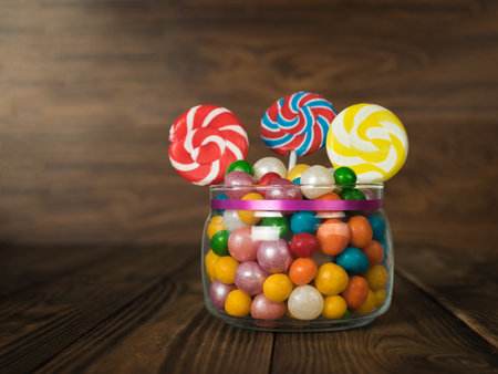 A glass jar with candy balls and lollipops on a stick on a wooden background. A treat for the party.