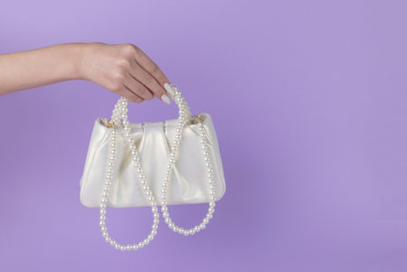 The girl's hand holds a white bag trimmed with pearls on a purple background. A fashionable women's accessory.の写真素材